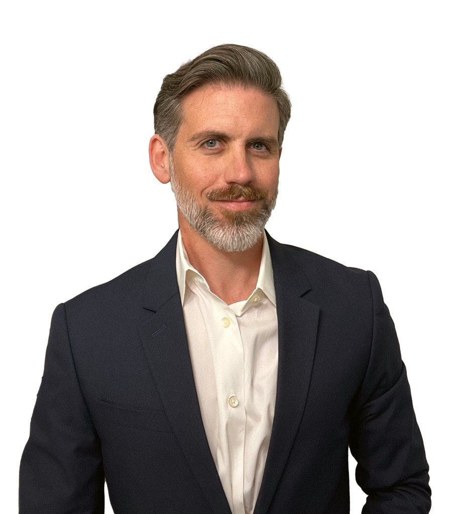 Photo of Tyler Hicks, President of Snakebite Consulting.  40 year old white male with patchy grey beard wearing white shirt and blue jacket